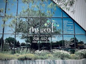 Front of a glass-walled wellness center with a large 'Answers' logo, a tree-and-question-mark graphic, a phone number, and landscaped plants in front.