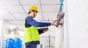 Maintenance worker wearing a yellow hard hat and high-visibility vest inspects a wall-mounted device, holding a tablet for reference